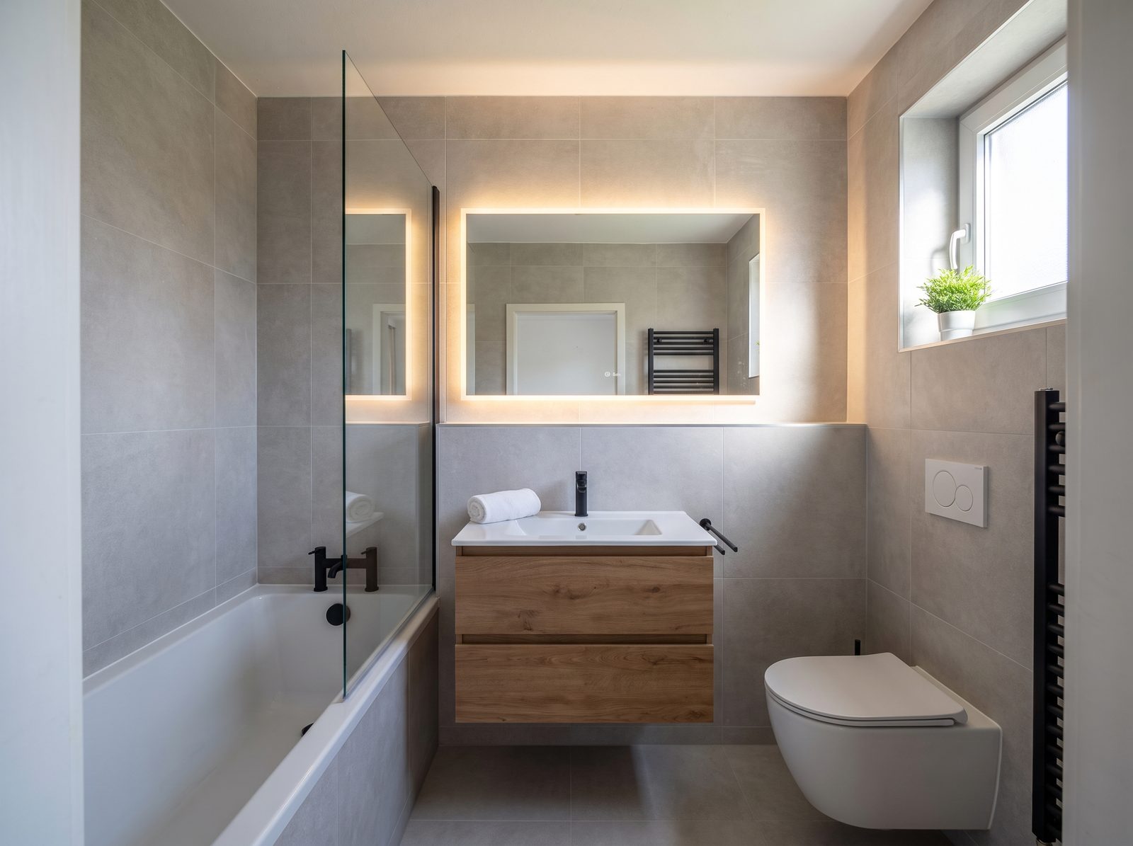 Dasselbe Badezimmer nach der Sanierung: gro&szlig;formatige helle Fliesen, Holz-Waschtisch mit beleuchtetem Spiegel, Badewanne mit Glasabtrennung und Wand-WC